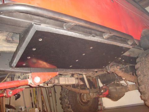 1989 Ford Bronco - Low-angle underbody view of a red 4x4 showing a black bolt-on skid plate/belly pan spanning the frame rails, tied into a tubular rock slide…