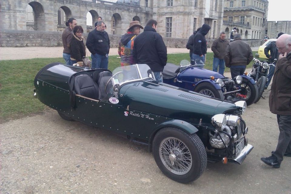 Several vintage three-wheeled sports cars parked outdoors with people standing around. The closest car is dark green with wire wheels and 'Rattle 'N Hum' on its side