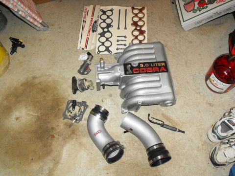 1994 Ford Bronco - A collection of engine parts, including a 5.0 Liter Cobra intake manifold, C&L intake tubes, and gaskets, laid out on a concrete floor.