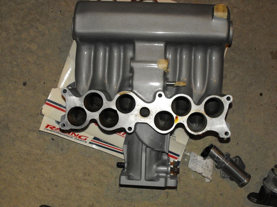 Silver intake manifold with eight intake ports, resting on packaging with 'RACING PERFORMANCE PARTS' text