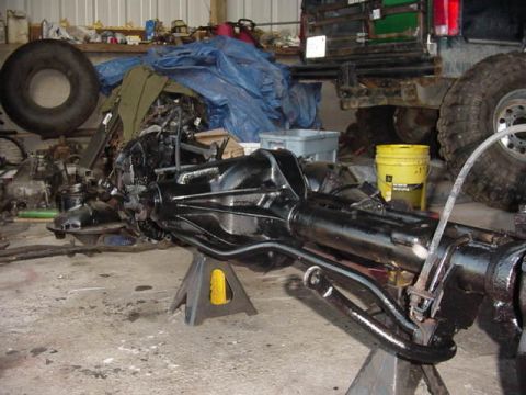 1992 Ford Bronco - A black front axle assembly is supported by jack stands in a garage setting, with a large tire and other vehicle parts in the background. T…