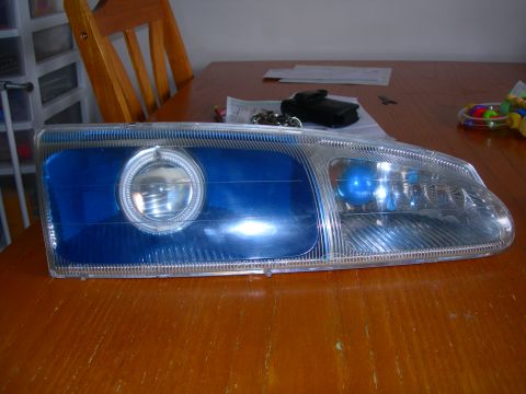 1984 Ford Bronco - A single headlight with a blue inner lens and a halo ring is displayed on a wooden table.