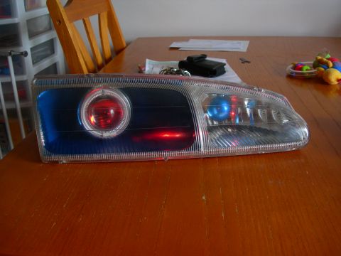 1984 Ford Bronco - Aftermarket headlight assembly with projector lens and red/blue LEDs on a wooden table.