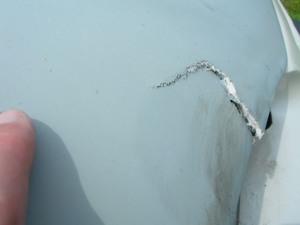 Close-up of a damaged light-colored vehicle panel with a torn section revealing underlying material. A thumb is visible on the left