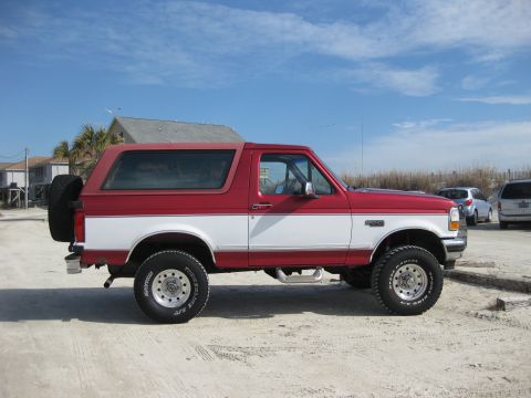 1995 Ford Bronco - Two-tone red and white 1995 Ford Bronco parked on a sandy lot near buildings under a bright blue sky.