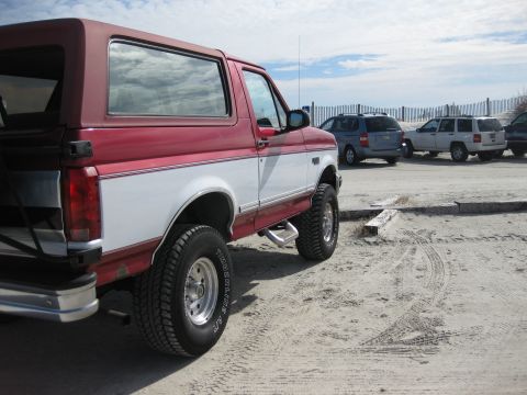 1995 Ford Bronco - This two-tone SUV is parked on a sandy lot near a wooden fence and other vehicles.