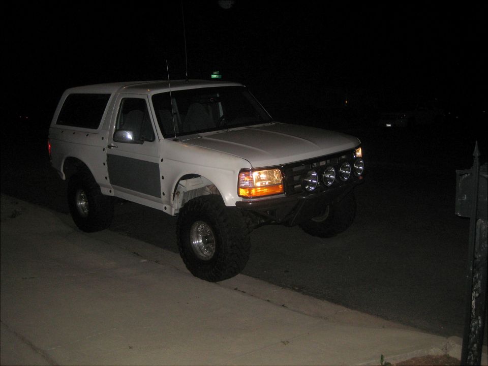 White 1992 Ford Bronco with aftermarket bumper and four fog lights, lifted suspension, and large tires, parked on a sidewalk at night