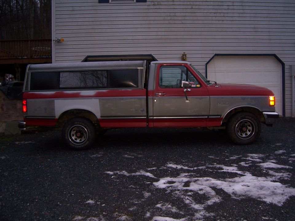 Side view of a red and gray pickup truck with a camper shell parked in front of a garage