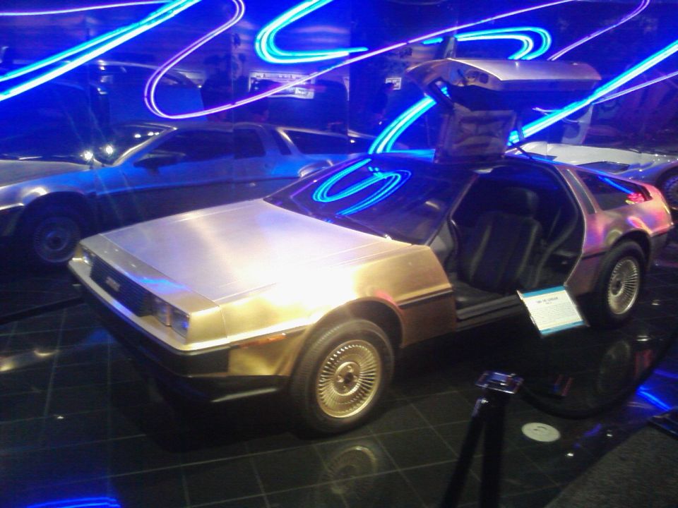 DeLorean DMC-12 with gullwing doors open, another DeLorean behind it, museum display with blue neon lights
