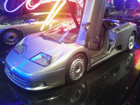 1995 Ford Taurus - A silver Bugatti EB110 is displayed with its doors open, illuminated by neon lights.