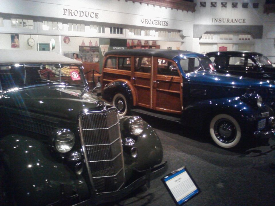 Museum exhibit with a dark green vintage car in the foreground and a blue woody wagon behind it