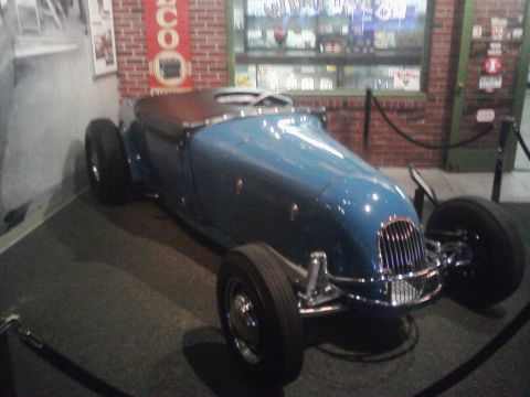 1995 Ford Taurus - A blue roadster with exposed front wheels and a chrome grille is displayed indoors.