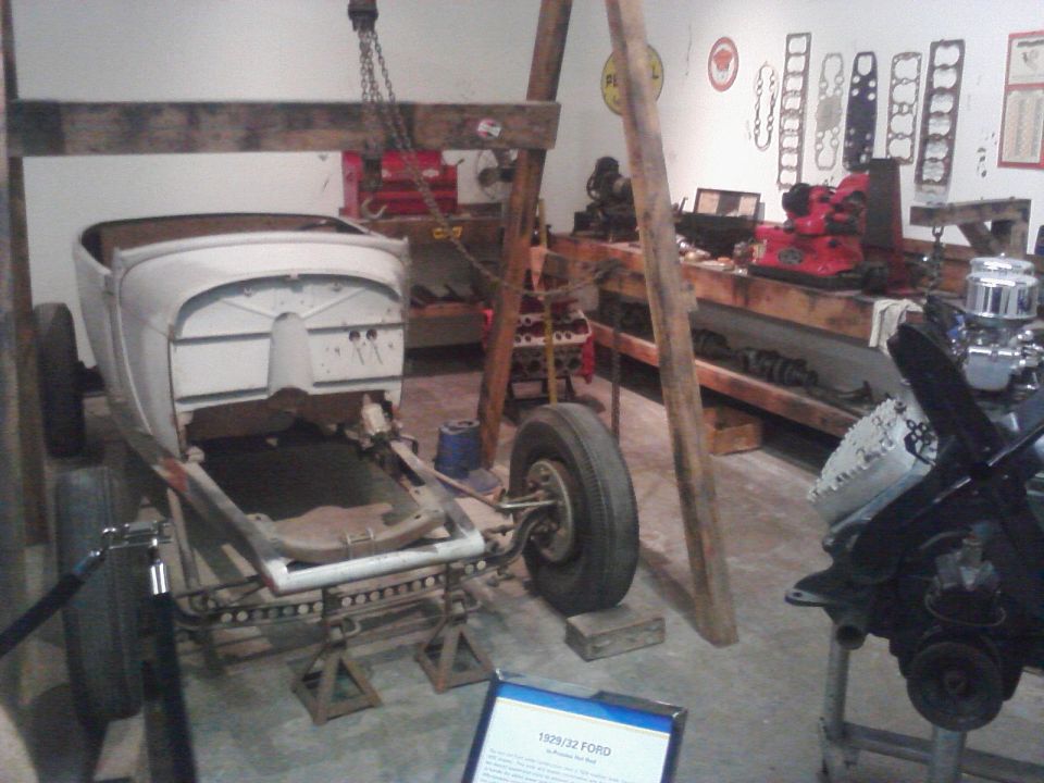 Vintage Ford hot rod body on a frame in a workshop with engine parts on benches, 1928-32 display sign