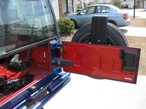 1998 Jeep Wrangler - The rear of a blue Jeep Wrangler is shown with its red tailgate open, revealing the cargo area and a spare tire. A blue sedan is parked nea…