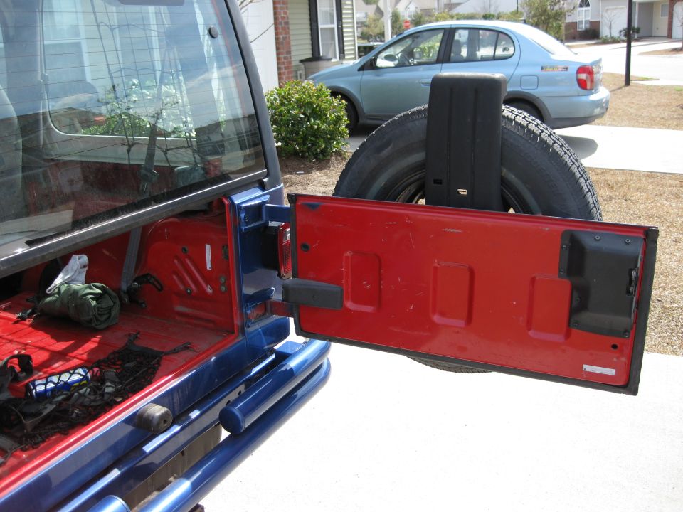Blue Jeep Wrangler rear with red tailgate open, spare tire mounted, and a blue sedan in the background