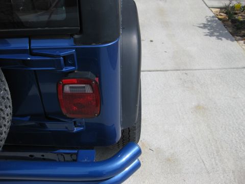 1998 Jeep Wrangler - The rear quarter panel of a blue Jeep Wrangler is visible, parked on a concrete driveway.