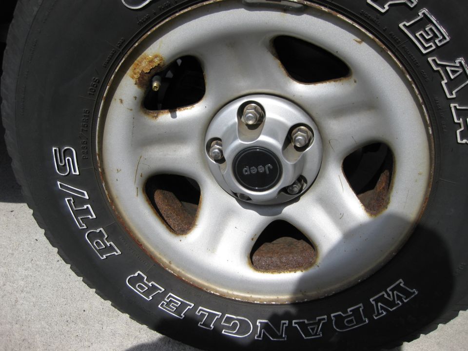 Close-up of a 1998 Jeep Wrangler wheel with a "Jeep" center cap and "Wrangler" tire sidewall. The wheel shows rust