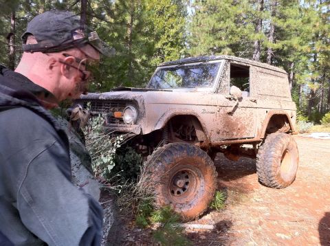 1989 Ford Bronco - A man inspects a heavily mud-caked classic Ford Bronco, likely after off-roading.