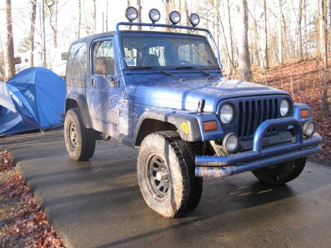 1998 Jeep Wrangler - A blue Jeep Wrangler with a flat tire and auxiliary lights is parked on a paved surface next to a tent in a wooded area.