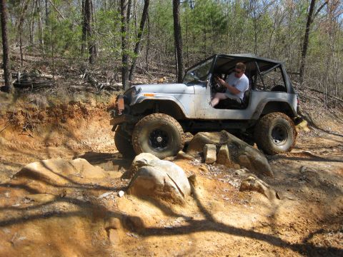 1998 Jeep Wrangler - A driver maneuvers a Jeep Wrangler over rough terrain on a dirt path surrounded by trees.