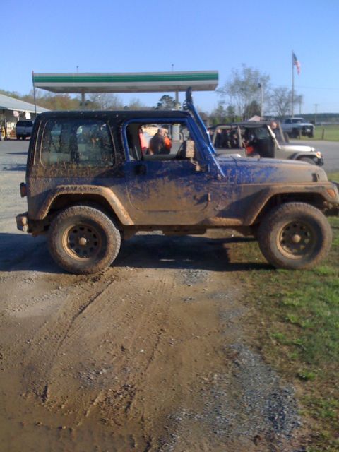 1998 Jeep Wrangler - This muddy blue Jeep Wrangler is seen from the side, with other vehicles and a gas station canopy in the background.