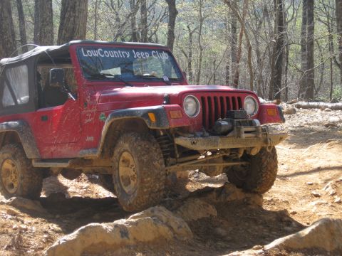 1998 Jeep Wrangler - A red Jeep Wrangler navigates a rocky trail in a wooded area, displaying a club decal on its windshield.