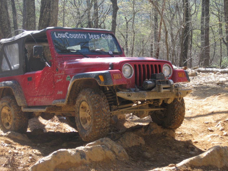 Red Jeep Wrangler with 'LowCountry Jeep Club' on windshield driving over rocks on a wooded trail