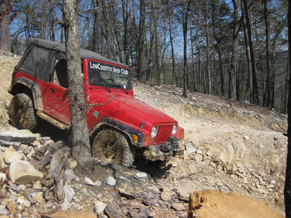 Red Jeep Wrangler with 'LowCountry Jeep Club' sticker on windshield, going over rocks on a wooded trail