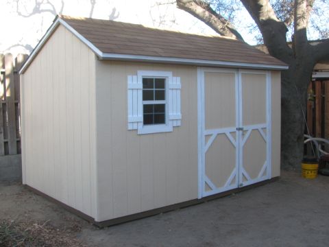 1968 Ford Mustang - Tan backyard storage shed with white trim, a shuttered window, brown shingled gable roof and white-braced double doors, standing on dirt be…