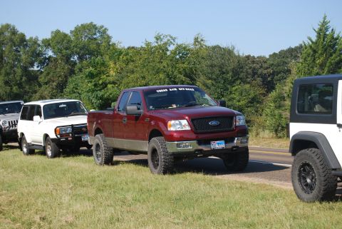 2005 Ford F-150 - A line of off-road vehicles, including a red Ford F-150, are parked on the side of a road under a clear sky.