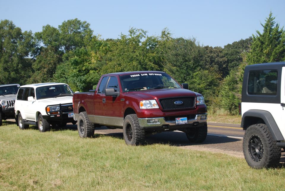 Red Ford F-150 pickup truck with lifted suspension and off-road tires, parked on grass next to a road. A "TEXAS LIL OFF ROAD" sticker is on the windshield