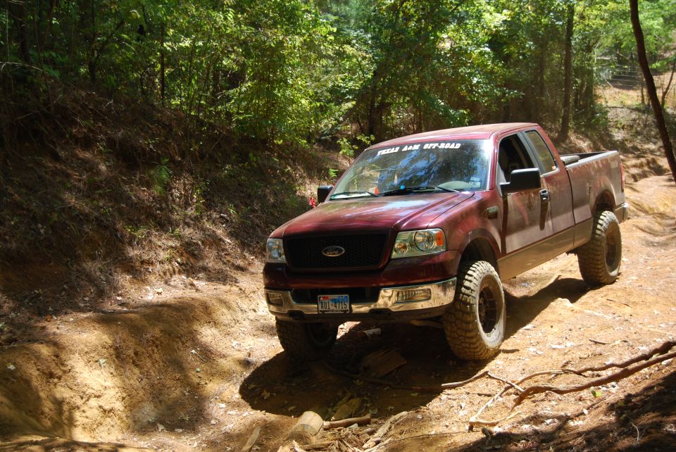 Maroon Ford F-150 driving on a dirt path in a wooded area. It has off-road tires and a sticker on the windshield that says 'TEXAS JAM OFF-ROAD'