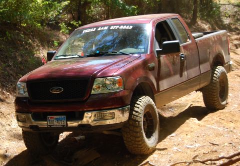 2005 Ford F-150 - A maroon Ford F-150 pickup truck with off-road tires drives on a dirt path surrounded by trees.