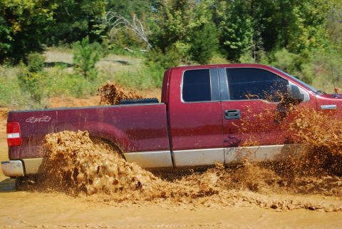 2005 Ford F-150 - A red 2005 Ford F-150 kicks up a large spray of mud as it drives through a muddy area.