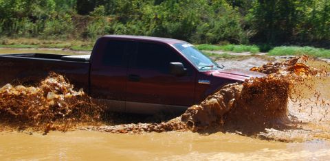 2005 Ford F-150 - A maroon 2005 Ford F-150 navigates a muddy off-road course, sending water and dirt flying.