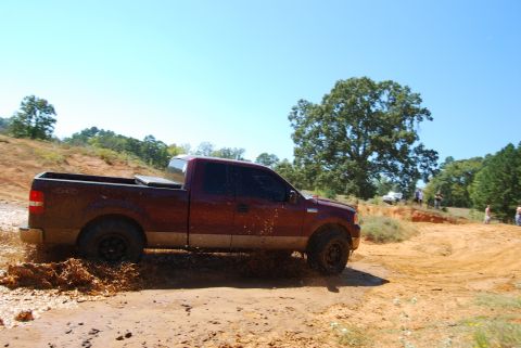 2005 Ford F-150 - A maroon pickup truck navigates a muddy off-road course with other vehicles and people in the background.