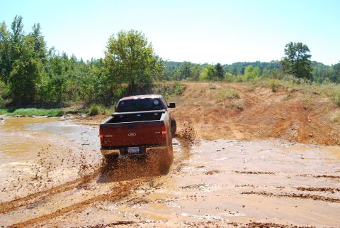 2005 Ford F-150 - A red 2005 Ford F-150 navigates a muddy off-road course, kicking up water and dirt.