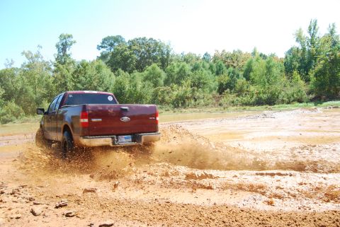 2005 Ford F-150 - A red pickup truck navigates a muddy terrain, creating a large splash as it moves forward.