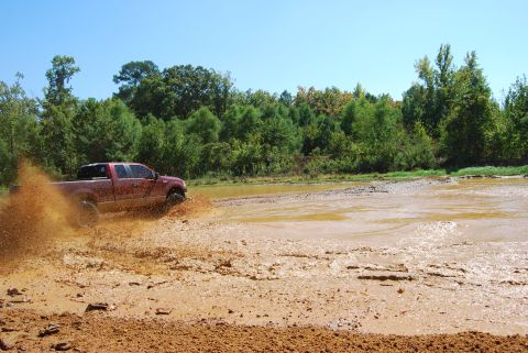 2005 Ford F-150 - A red 2005 Ford F-150 navigates a muddy terrain, kicking up a large spray of water and dirt.