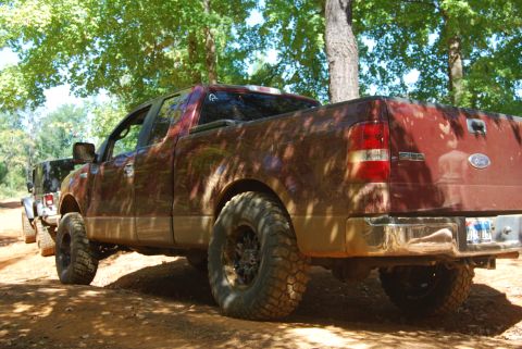 2005 Ford F-150 - A red 2005 Ford F-150 pickup truck with large off-road tires is seen from the rear 3/4 angle, parked on a dirt road.