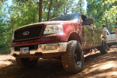 2005 Ford F-150 - Muddy red Ford F-150 with aftermarket wheels and tires parked on a dirt path with trees in the background.