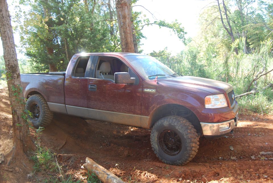 Maroon Ford F-150 pickup truck with lifted suspension and off-road tires driving on a dirt trail