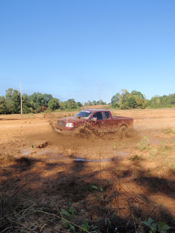 Red pickup truck splashing mud while driving through a muddy field. The truck appears to be from the early 2000s