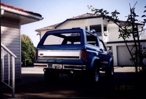 1985 Ford Bronco - Rear view of a blue 1985 Ford Bronco with a silver stripe, parked in a residential driveway.