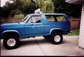 1985 Ford Bronco - Side view of a blue SUV with white rims parked outdoors.
