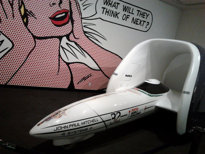 streamliner vehicle with 'JOHN PAUL MITCHELL SYSTEMS' and 'PENTAX WORLD LAND SPEED CHALLENGE' text, next to pop art