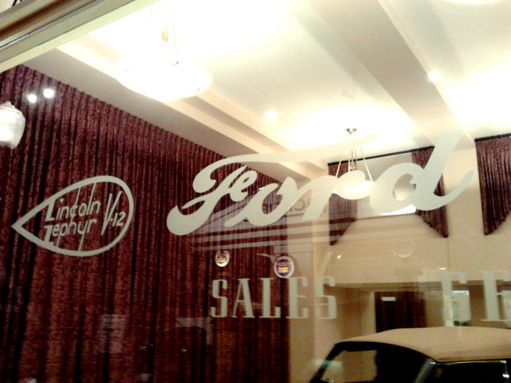 Window of a Ford dealership with 'Ford' and 'Lincoln Zephyr V12' logos etched on it. 'SALES' is also visible