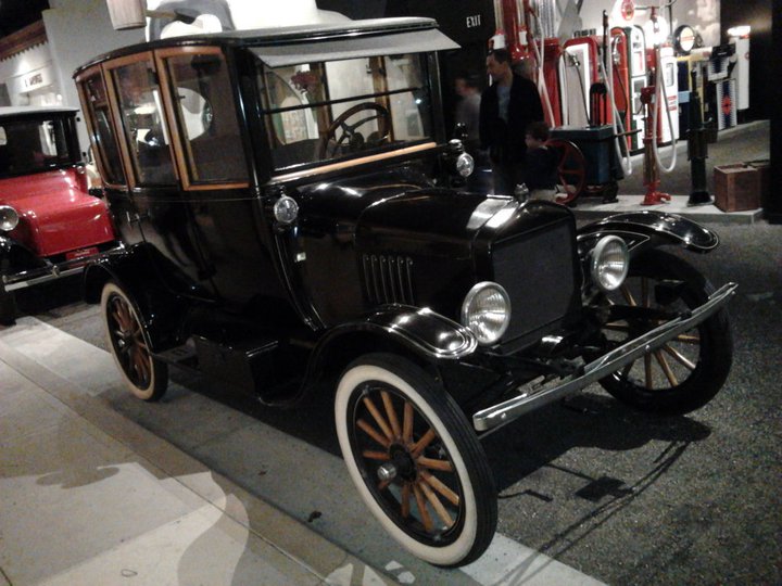Black antique touring car with wooden window frames and whitewall tires. Red car visible behind it. Gas pumps in background