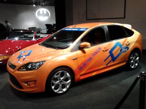 1995 Ford Taurus - An orange Ford Focus electric car is displayed at the Peterson Automotive Museum, next to a red convertible.
