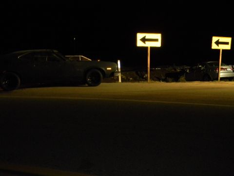 1975 Ford Ranchero - A dark sedan with black wheels is parked at night next to a road with directional signs.